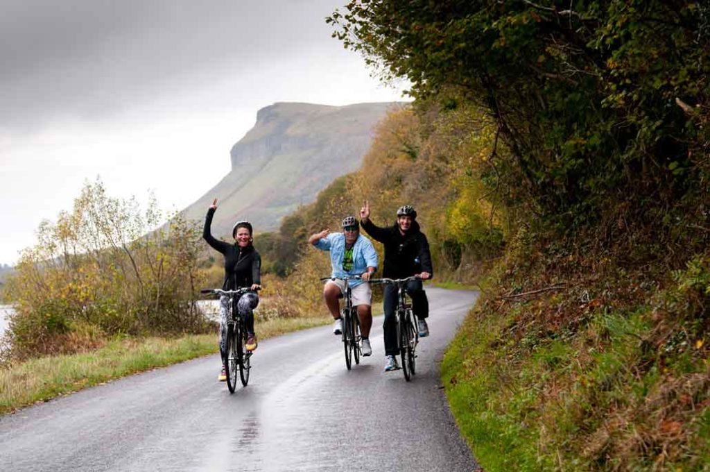 Three people cycling on road sticking their hands up to wave at cameraman